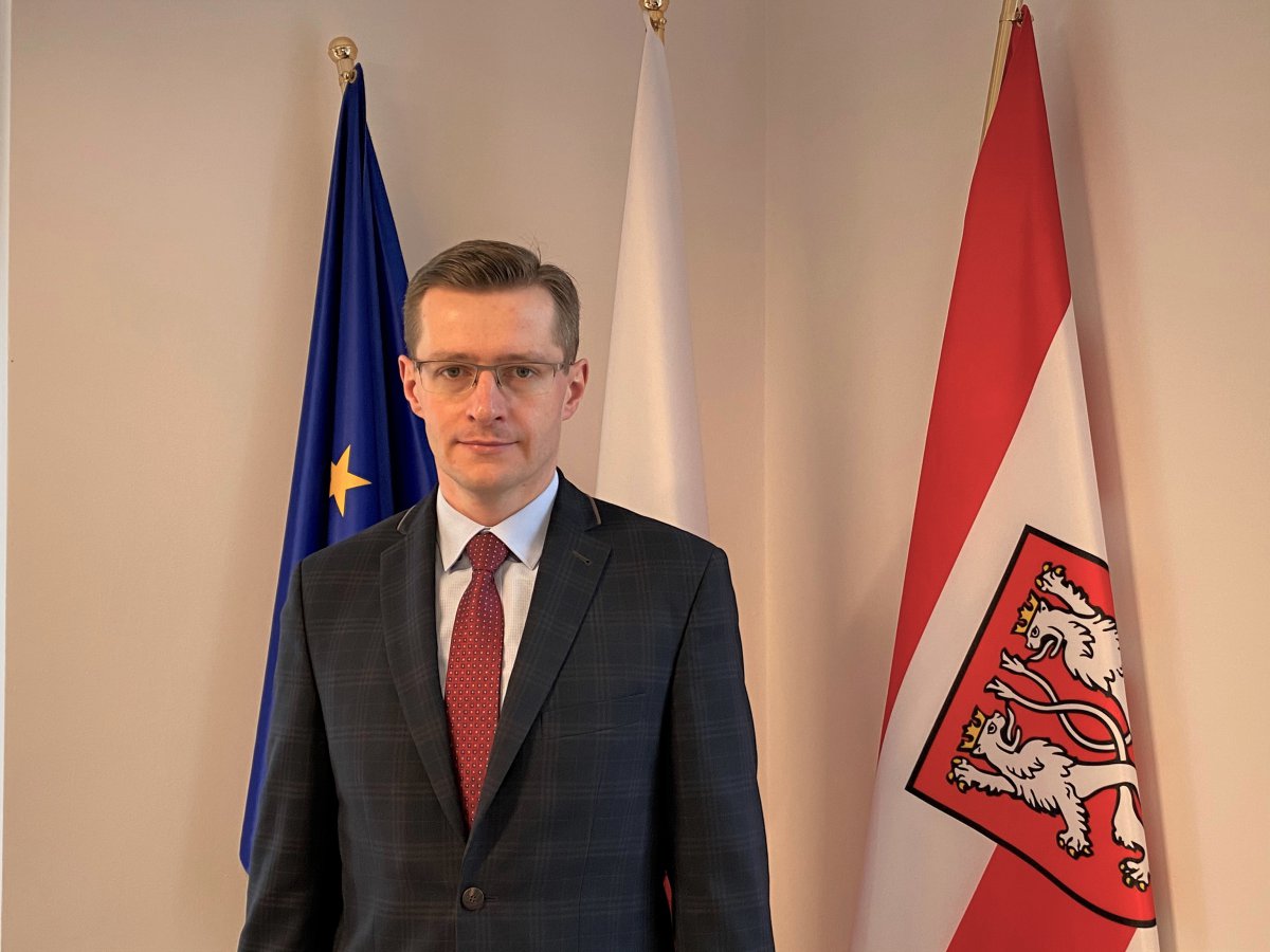 mężczyzna stojący przed trzema flagami: europejską, polską i gminną