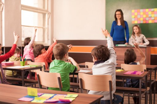 zdjęcie przedstawia salę lekcyjną w której siedzą dzieci z uniesionymi do góry rękami, przed tablica stoi nauczycielka z jedną uczennicą